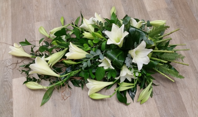 White flower and foliage single ended funeral coffin spray with longi lilies and stems at the end to look like a hand-tied arrangement