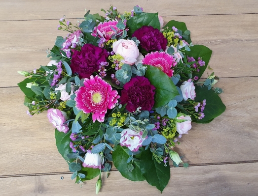 A round funeral flower posy with rich plum carnations, bright pink gerberas, and soft pink roses, accented with seasonal blooms and lush greenery.