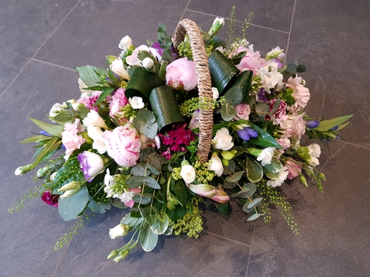 Funeral basket arrangement with pink peonies, pink roses, baby pink carnations, scented white freesia, and blue-lilac accents.