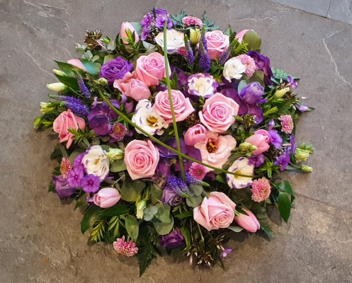 A round funeral posy with soft pink roses, delicate purple lisianthus, and seasonal blooms in shades of pink, purple and lilac.