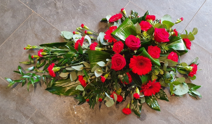 A single ended funeral spray with red roses, bold red gerberas, and charming red carnations, complemented by glossy tropical foliage and soft greenery