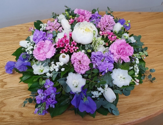 A round funeral flower arrangement featuring scented lilac stocks, white and purples lisianthus, and pink carnations. Accented with seasonal flowers and lush greenery.