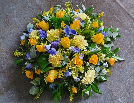 A round funeral posy with vibrant yellow roses, delicate blue delphiniums, and soft lemon carnations. Accented with seasonal blooms and lush greenery,