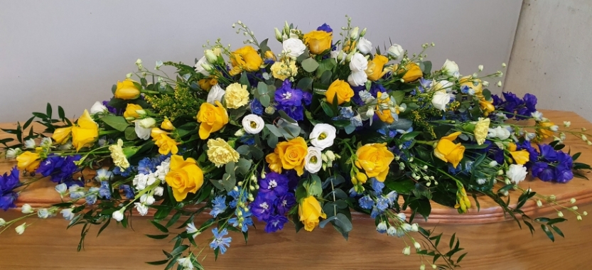 Coffin spray with golden yellow roses, blue delphiniums, velvet purples, and pure whites, with mixed foliage.