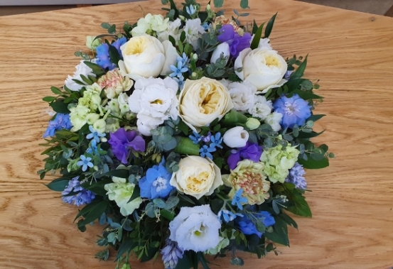 Round funeral posy with ivory roses, blue delphiniums, and seasonal blooms in shades of blue, purple, and green.