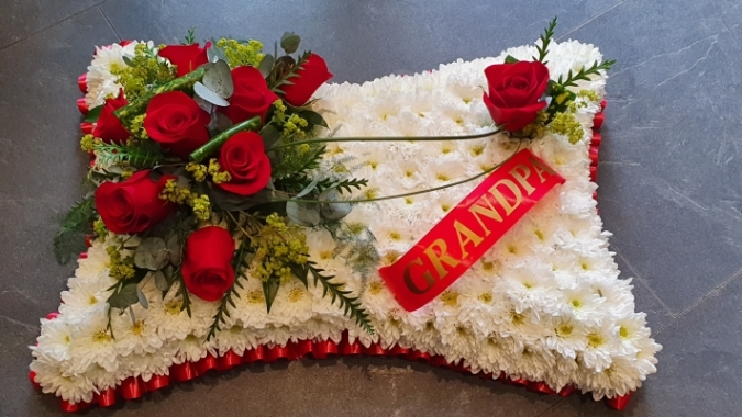 A pillow-shaped tribute based in pure white chrysanthemums, with a red rose spray, finished with a bold red ribbon edge and the option for a personalised name ribbon.