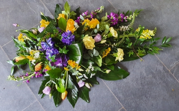 Single ended funeral spray with blue hyacinth, strong yellow roses, scented stocks, yellow antirrhinum, tulips and lisianthus with glossy foliage.