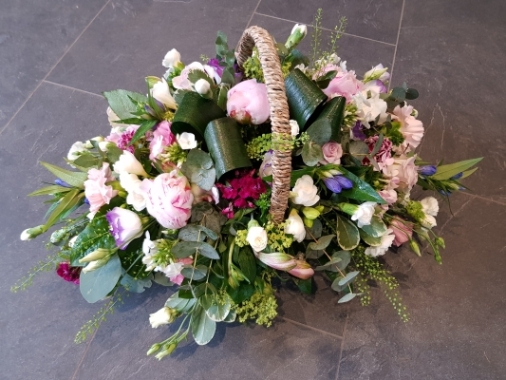 Funeral basket arrangement with pink peonies, pink roses, baby pink carnations, scented white freesia, and blue-lilac accents.