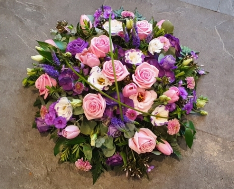 A round funeral posy with soft pink roses, delicate purple lisianthus, and seasonal blooms in shades of pink, purple and lilac.