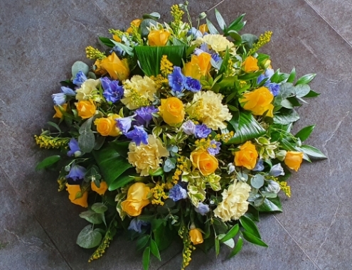 A round funeral posy with vibrant yellow roses, delicate blue delphiniums, and soft lemon carnations. Accented with seasonal blooms and lush greenery,